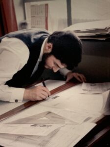 A color photograph of H.J. (John) Jackson leaning over a large desk, focused on drawing or writing with a pen. The desk is covered with technical drawings, papers, and a triangular ruler. He is wearing a white shirt and dark vest, and the background includes a window, stacked documents, and a wall calendar.