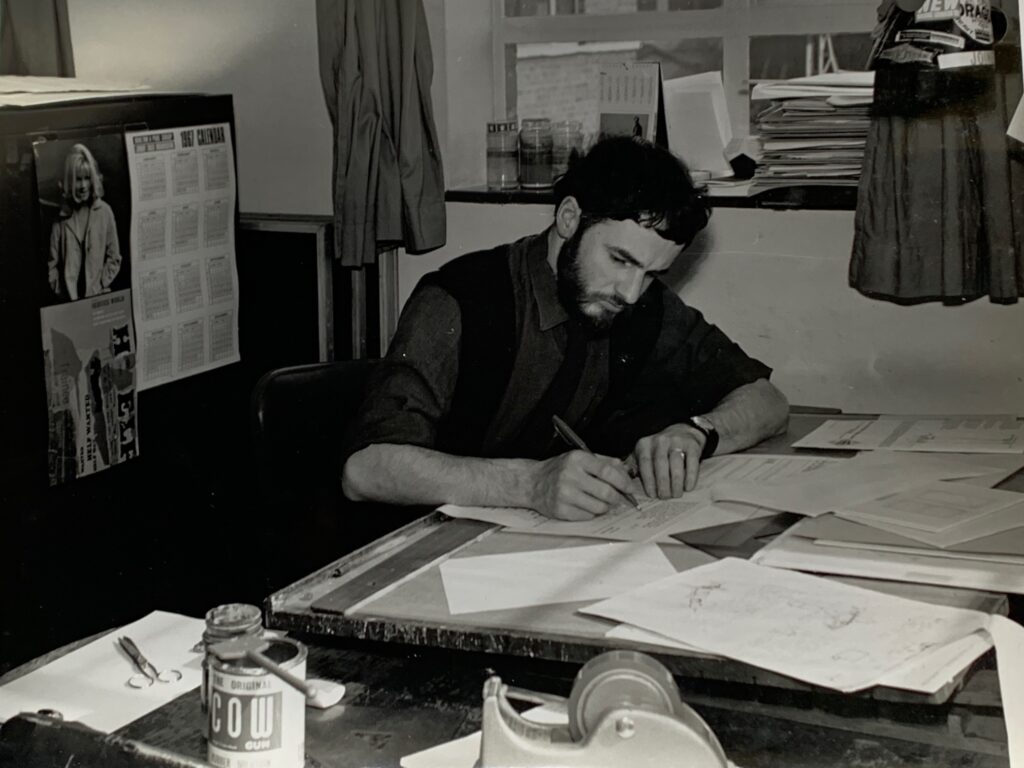 A black-and-white photograph of H.J. (John) Jackson sitting at a desk, writing on a piece of paper. The desk is cluttered with papers, a container labeled 'Cow Gum,' scissors, and other office supplies. In the background, there is a coat hanging, a calendar on the wall, and stacks of documents.