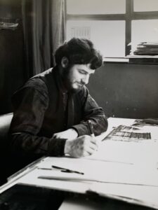 A black-and-white photograph of H.J. (John) Jackson with dark hair and a beard sitting at a desk, focused on drawing. The desk is covered with papers, including what appears to be a photograph or technical drawing. He is wearing a dark vest and shirt. The background includes a window with stacks of documents on the sill and a partially visible calendar on the wall.