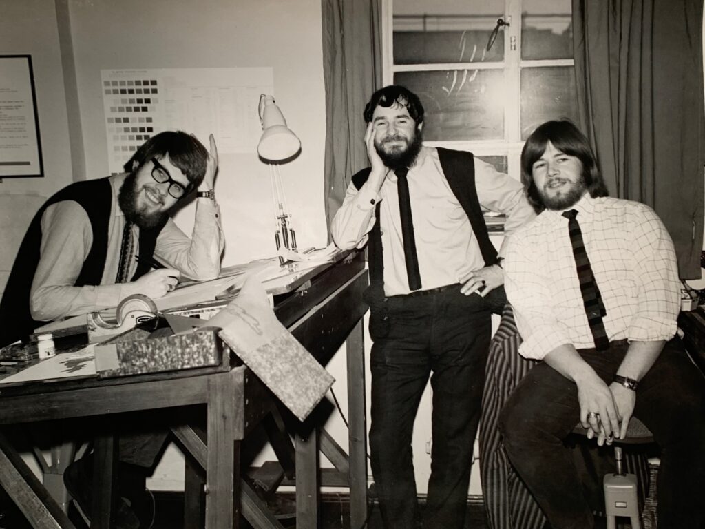 A black-and-white photograph featuring three men. Andrew Lear is seated at a drawing table. Beside him, H.J. (John) Jackson and Ian Kerridge pose for the photograph. The setting appears to be a workspace with drafting tools and materials visible in the background.