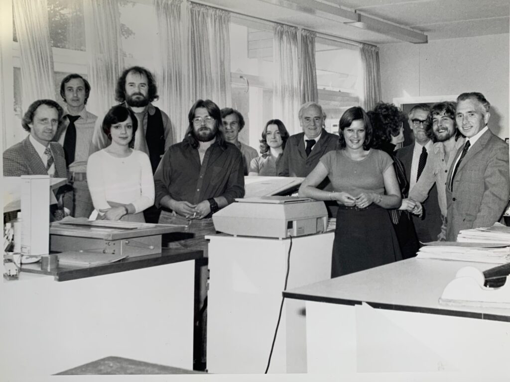 A black-and-white photograph of thirteen people posed in an office setting. A weighing scale is visible in the foreground. The room appears bright, with open windows in the background allowing natural light to fill the space.