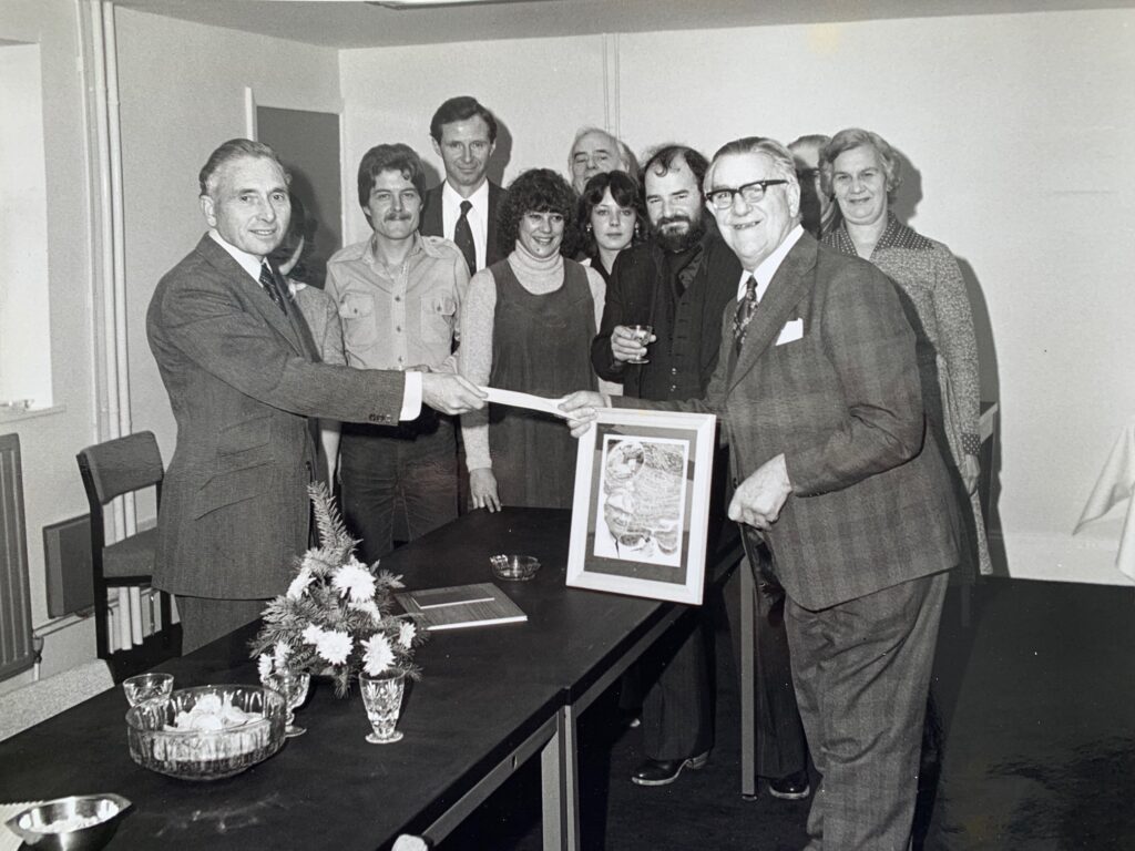 A black-and-white group photograph of eleven people gathered in a room. A long table in the foreground is decorated with flowers. Jack Paul is at the center of attention, being presented with framed artwork by H.J. Jackson.