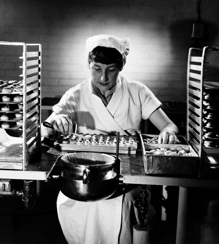 Person wearing a white uniform and cap, seated at a worktable, sorting chocolates on a conveyor with trays, in a production setting.