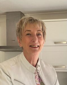 A cheerful older woman with short blonde hair smiles warmly at the camera. She is wearing a light-coloured jacket with a floral-patterned shirt underneath. 